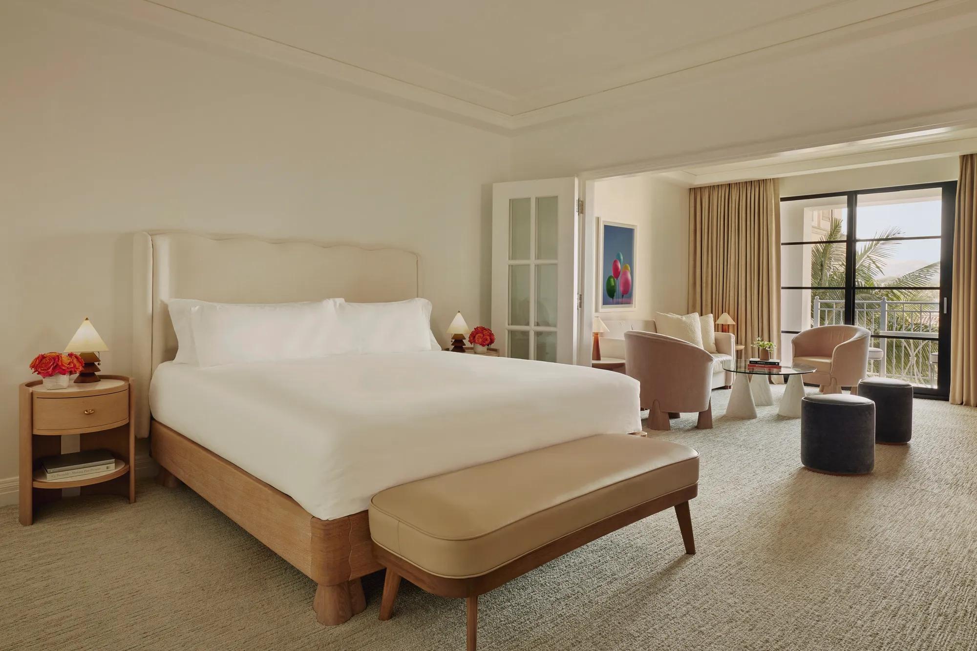 Junior suite with king bed and bench leading to a separate sitting area through French doors, opening to a balcony with palm views.