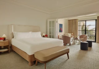 Junior suite with king bed and bench leading to a separate sitting area through French doors, opening to a balcony with palm views.