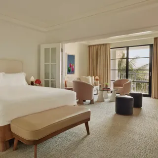 Junior suite with king bed and bench leading to a separate sitting area through French doors, opening to a balcony with palm views.