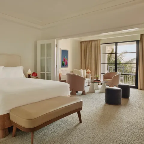 Junior suite with king bed and bench leading to a separate sitting area through French doors, opening to a balcony with palm views.