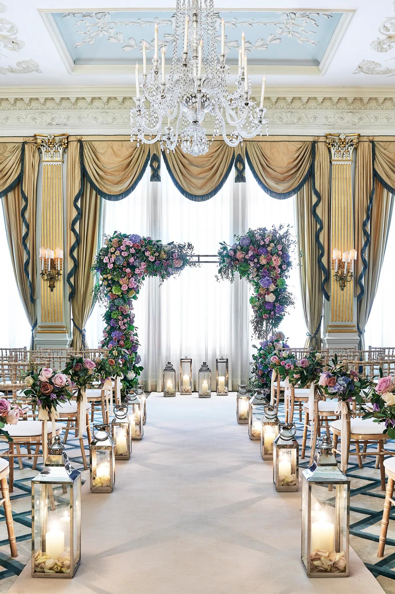 Elegant ballroom wedding ceremony setup with rows of gold chairs, floral arches of pink and purple blooms, tall lanterns lining the aisle, and a crystal chandelier hanging above in a room draped with gold and blue curtains.