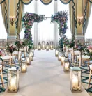 Elegant ballroom wedding ceremony setup with rows of gold chairs, floral arches of pink and purple blooms, tall lanterns lining the aisle, and a crystal chandelier hanging above in a room draped with gold and blue curtains.