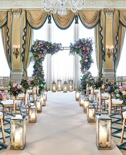Elegant ballroom wedding ceremony setup with rows of gold chairs, floral arches of pink and purple blooms, tall lanterns lining the aisle, and a crystal chandelier hanging above in a room draped with gold and blue curtains.