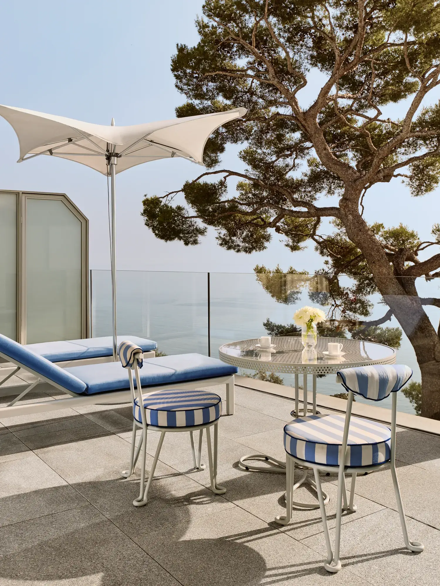 Terrasse avec chaises rayées et table ronde dressée pour le café, accompagnée d’un transat sous un parasol blanc, donnant sur la mer et encadrée par un grand arbre.