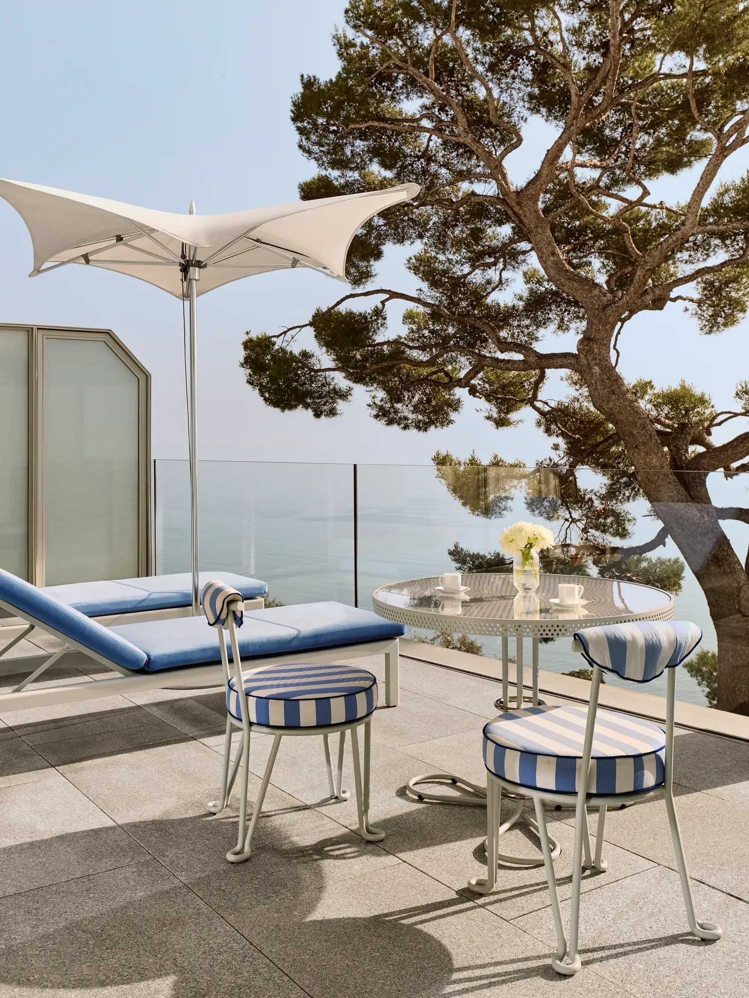 Terrasse avec chaises rayées et table ronde dressée pour le café, accompagnée d’un transat sous un parasol blanc, donnant sur la mer et encadrée par un grand arbre.