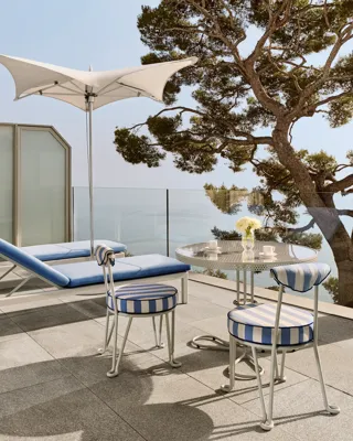 Terrasse avec chaises rayées et table ronde dressée pour le café, accompagnée d’un transat sous un parasol blanc, donnant sur la mer et encadrée par un grand arbre.