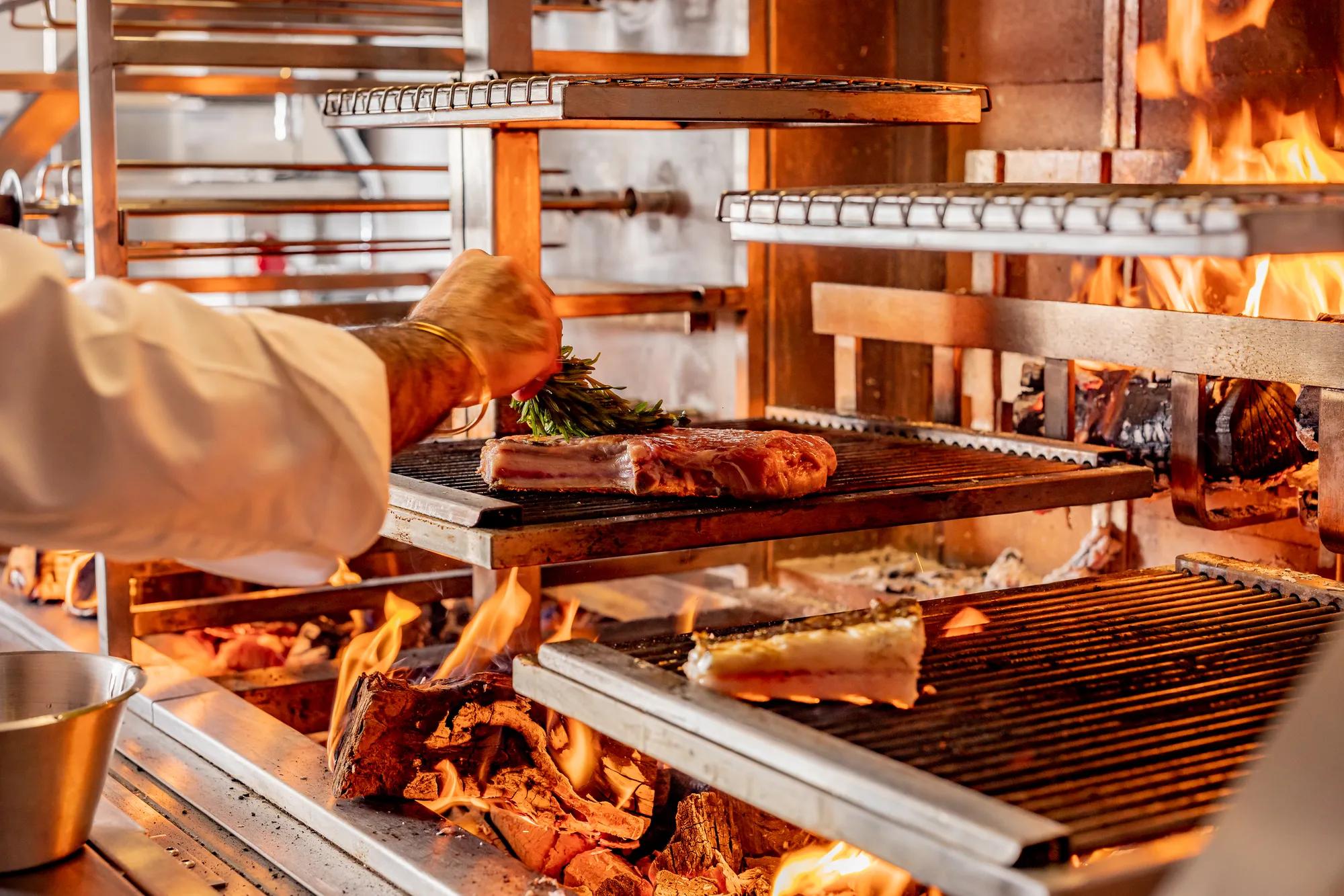 Chef placing herbs on meat cooking over open flames on a multi-level wood-fired grill.