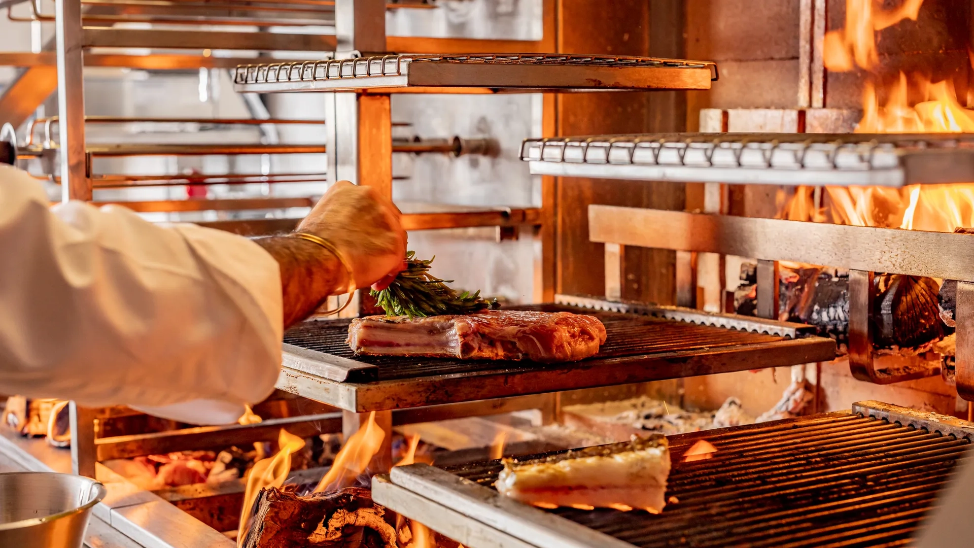 Chef placing herbs on meat cooking over open flames on a multi-level wood-fired grill.