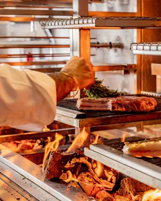 Chef placing herbs on meat cooking over open flames on a multi-level wood-fired grill.