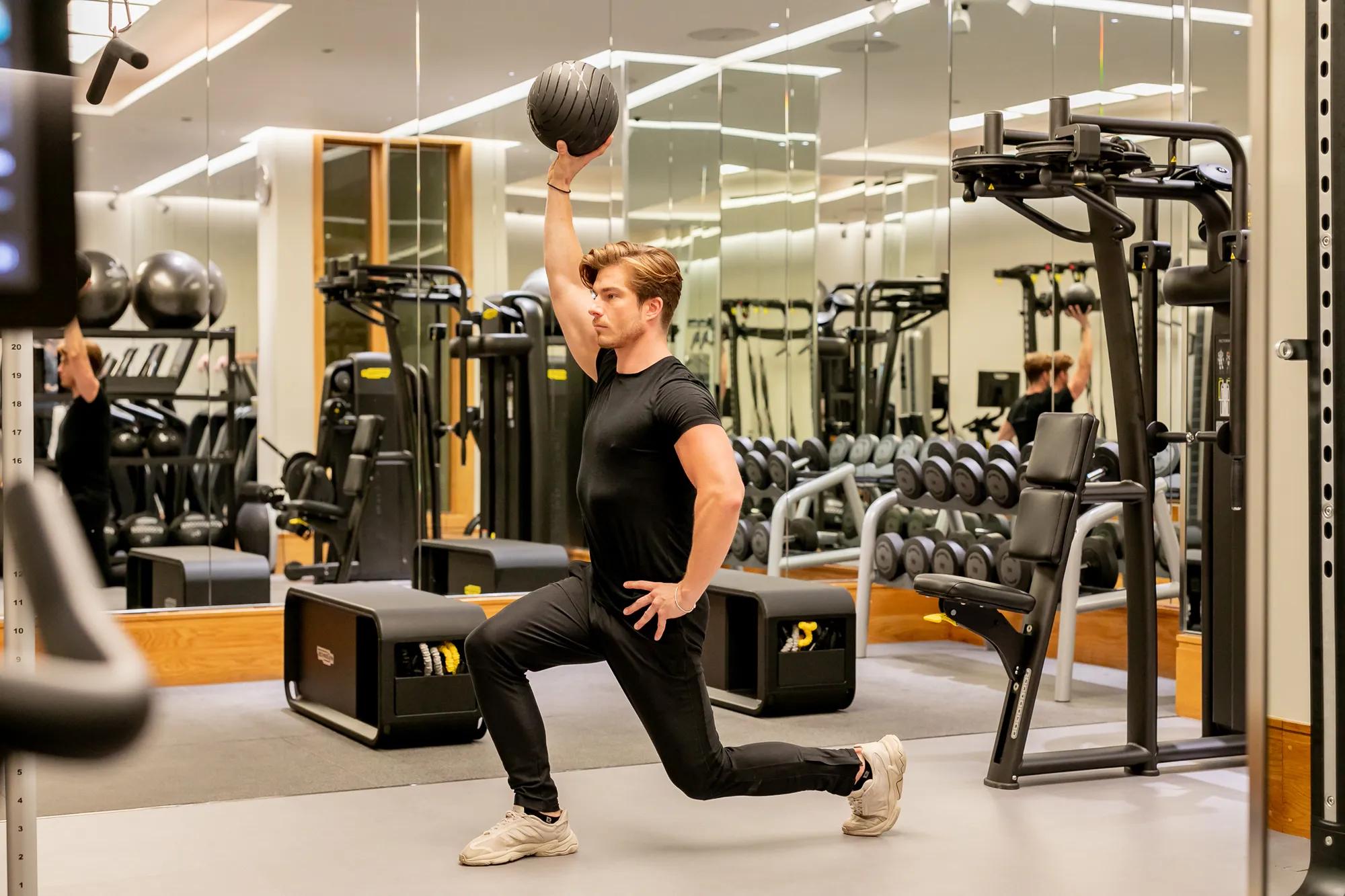 Man lunging with one knee on the floor in a mirrored gym, lifting a medicine ball overhead during a strength workout.