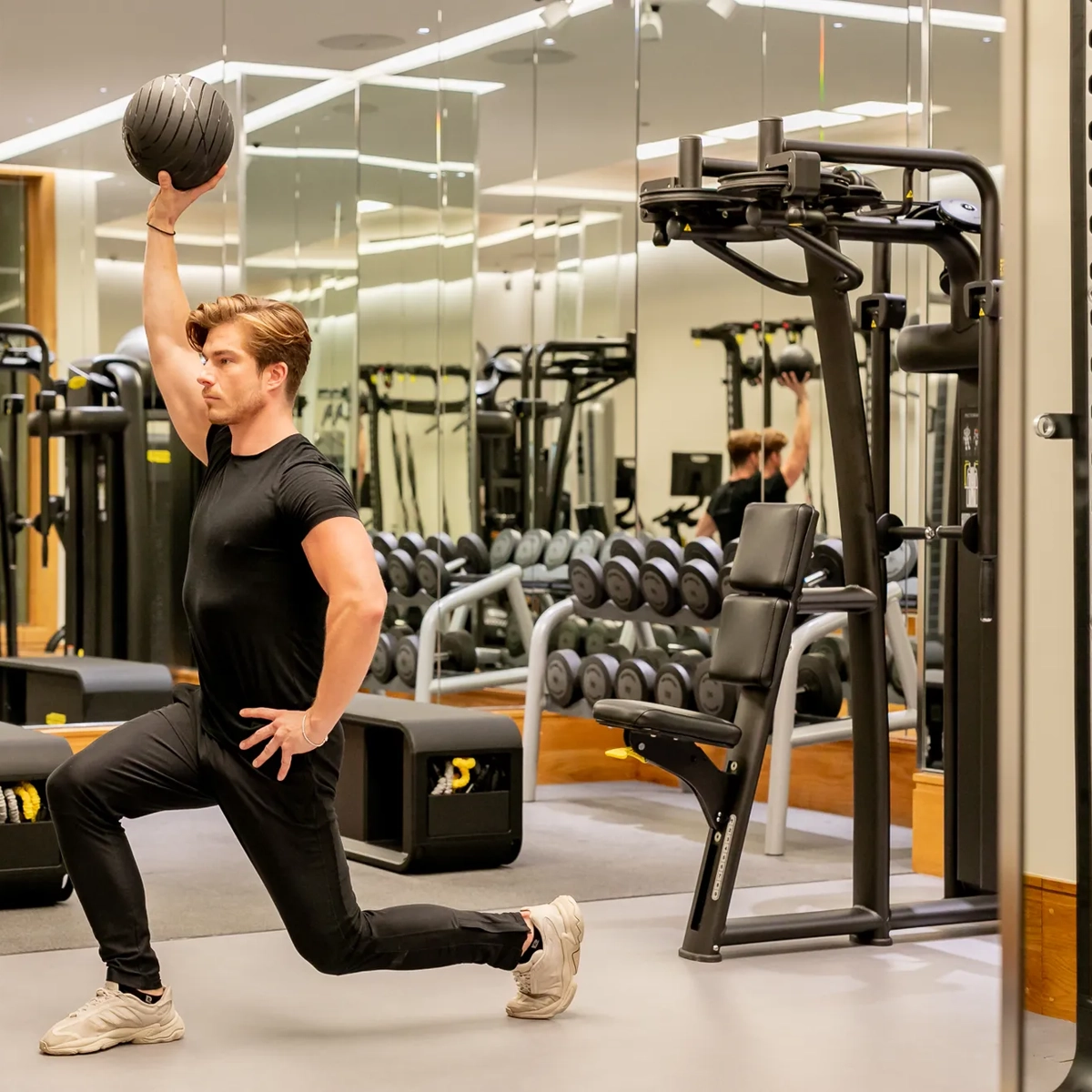 Man lunging with one knee on the floor in a mirrored gym, lifting a medicine ball overhead during a strength workout.