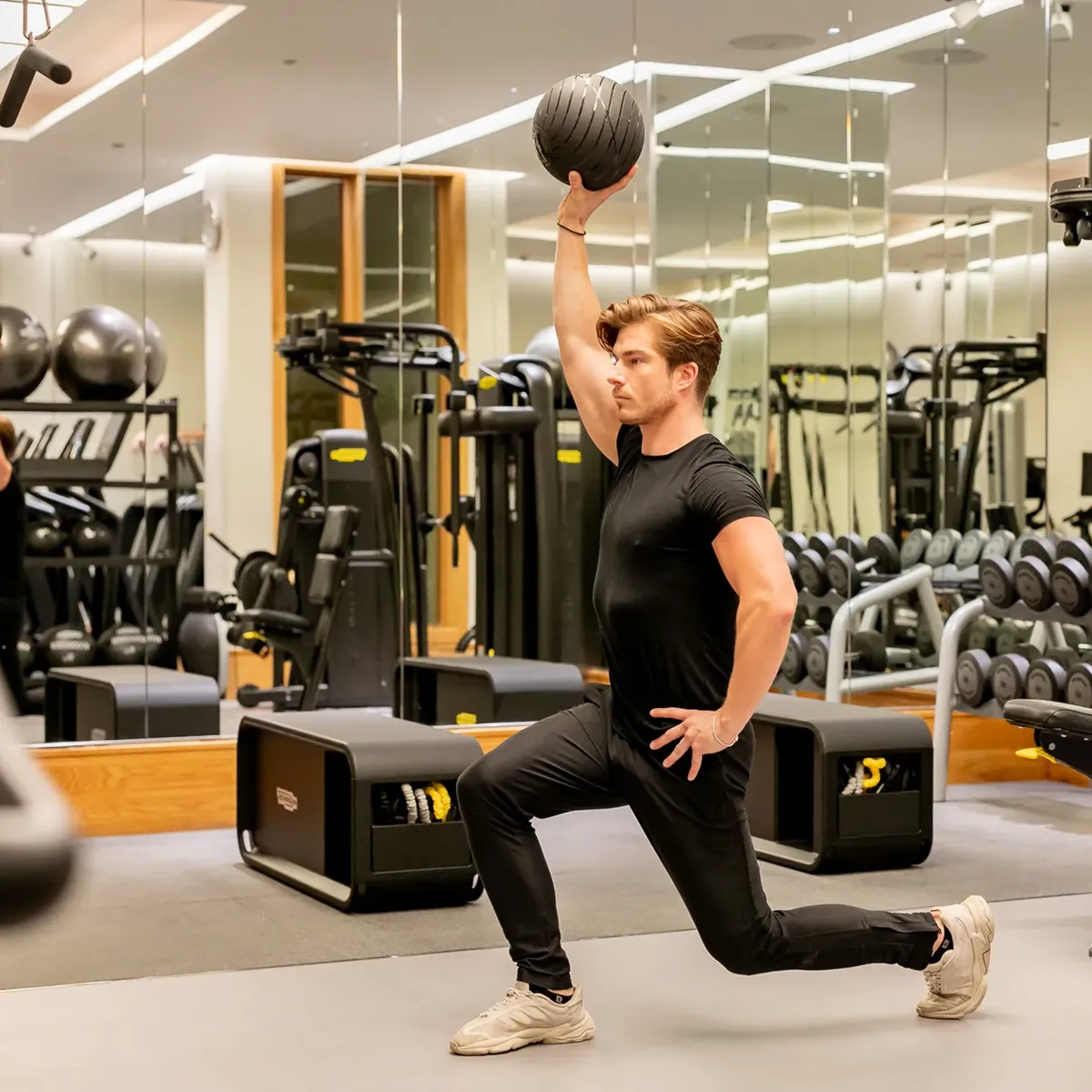 Man lunging with one knee on the floor in a mirrored gym, lifting a medicine ball overhead during a strength workout.