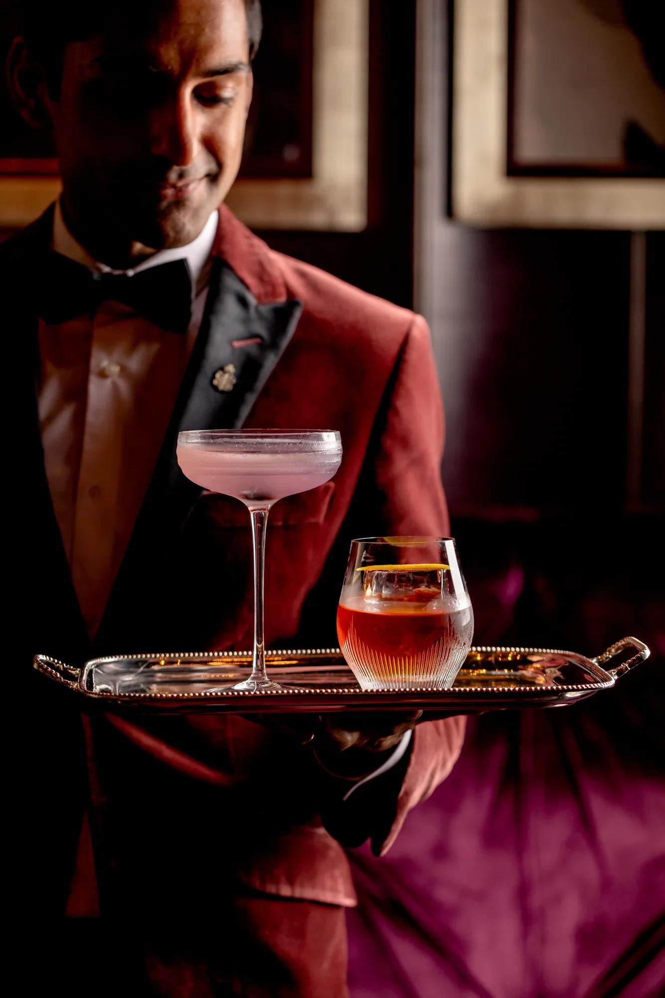 Server in a burgundy jacket presenting a tray with two elegant cocktails in a softly lit lounge.