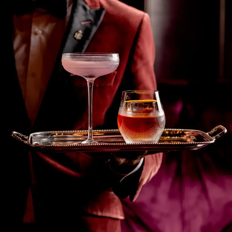 Server in a burgundy jacket presenting a tray with two elegant cocktails in a softly lit lounge.