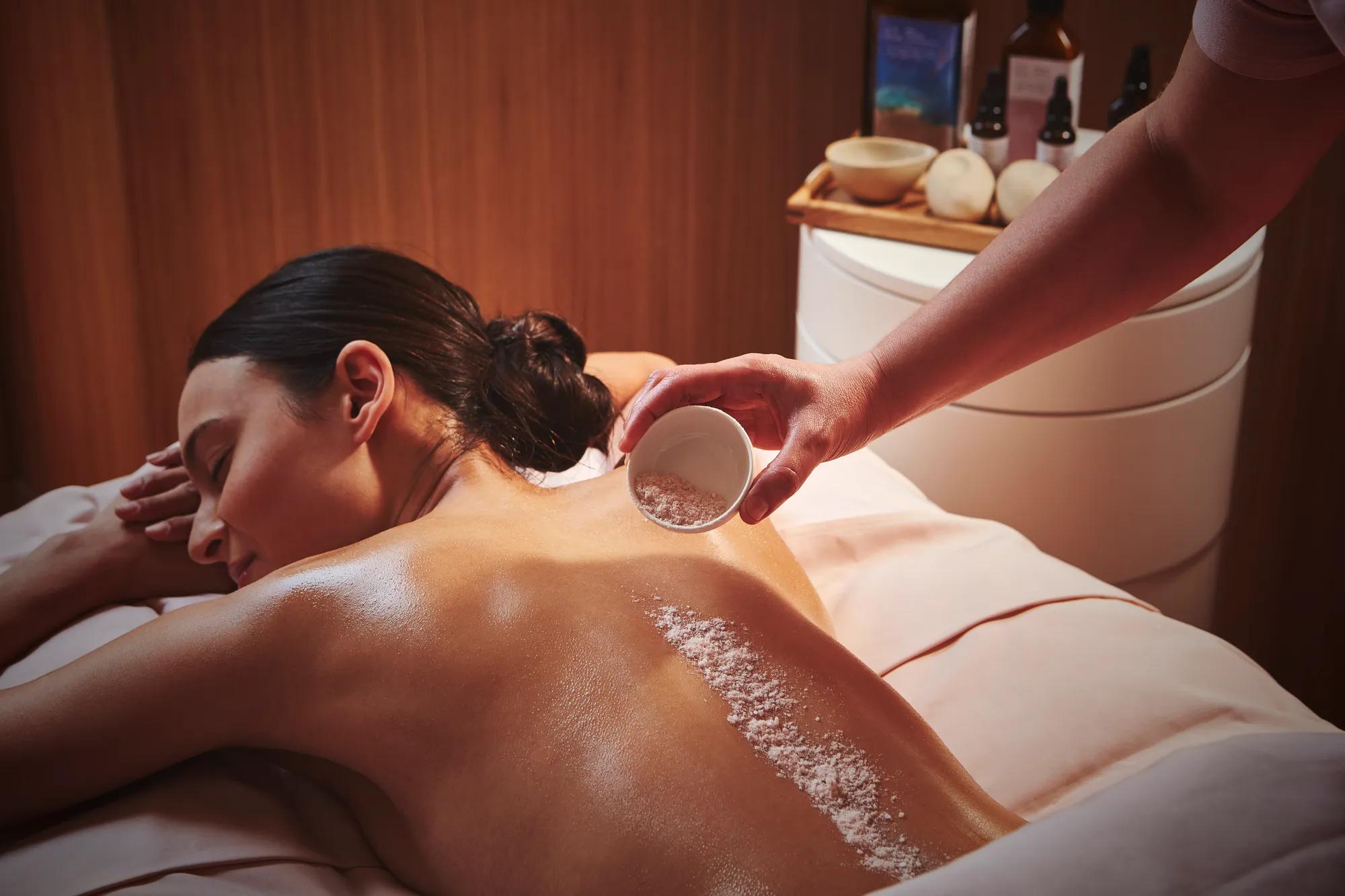 A spa therapist applies sea salt to a woman’s back during an exfoliating body treatment in a softly lit therapy room at The Maybourne Riviera.