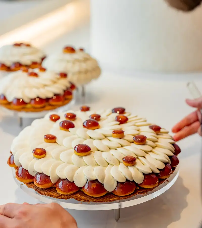 A pastry chef adds finishing touches to a Cédric Grolet tart topped with piped cream petals and glossy caramelised domes.