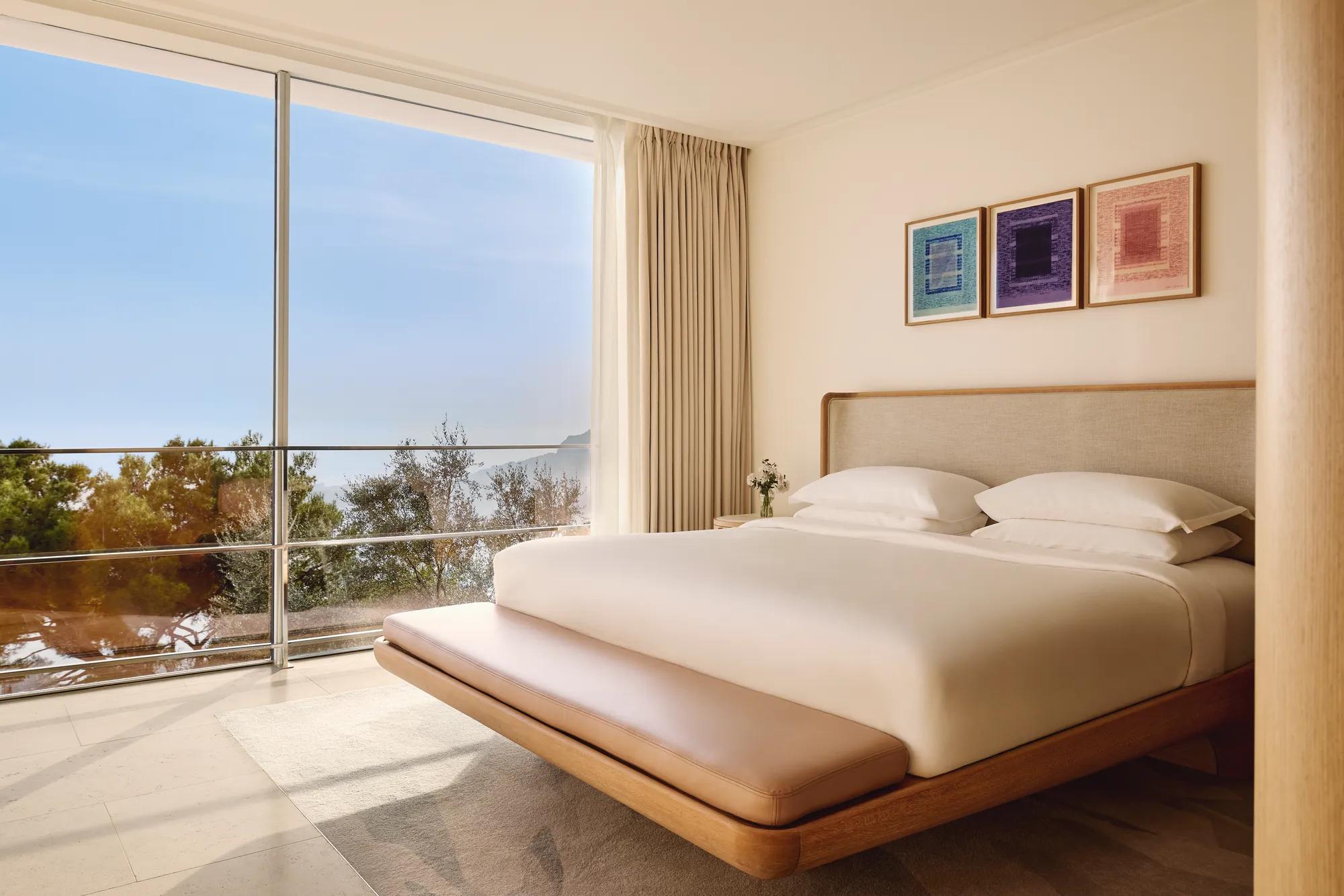 Elegant bedroom with a modern wooden bed, crisp white linens, and three colorful framed artworks, featuring floor-to-ceiling windows overlooking the sea and trees.