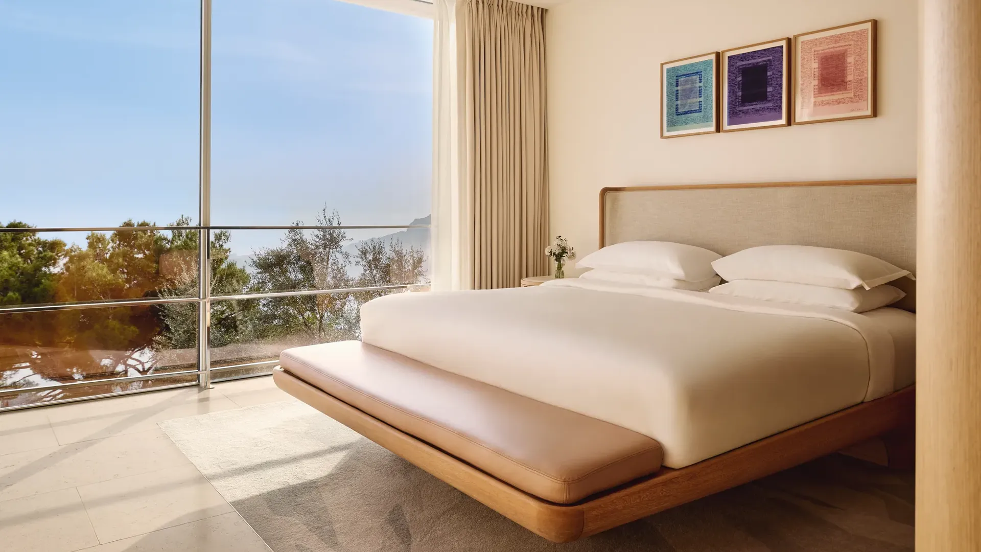 Elegant bedroom with a modern wooden bed, crisp white linens, and three colorful framed artworks, featuring floor-to-ceiling windows overlooking the sea and trees.