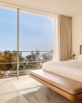 Chambre élégante avec lit moderne en bois, linge blanc immaculé et trois œuvres encadrées colorées, dotée de baies vitrées donnant sur la mer et les arbres.