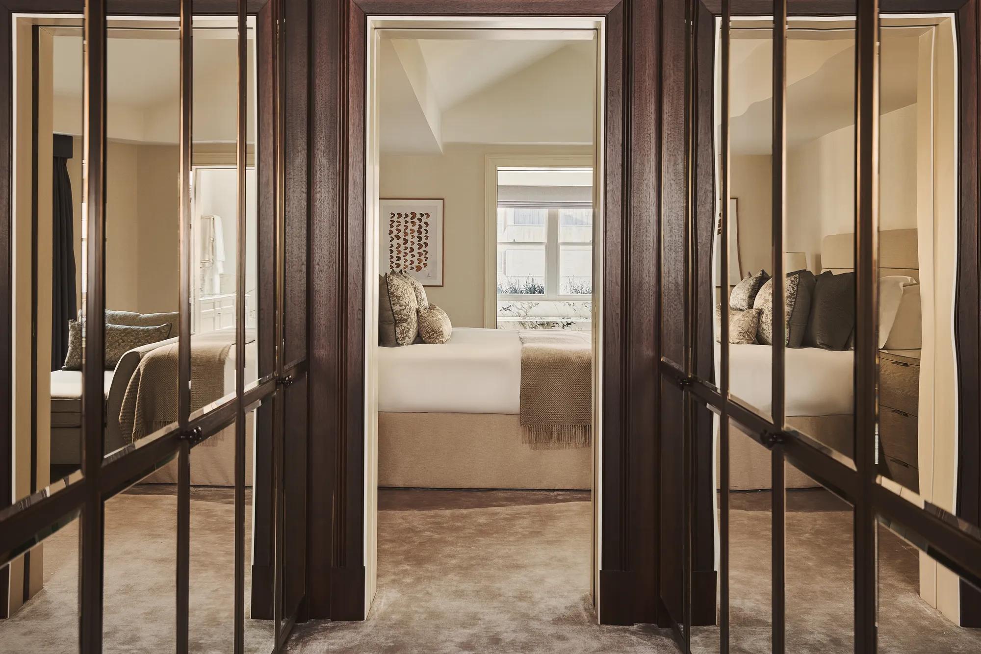 View through mirrored wardrobe doors towards a neutral-toned bedroom with a neatly made bed, soft lighting, and window bringing in natural daylight.