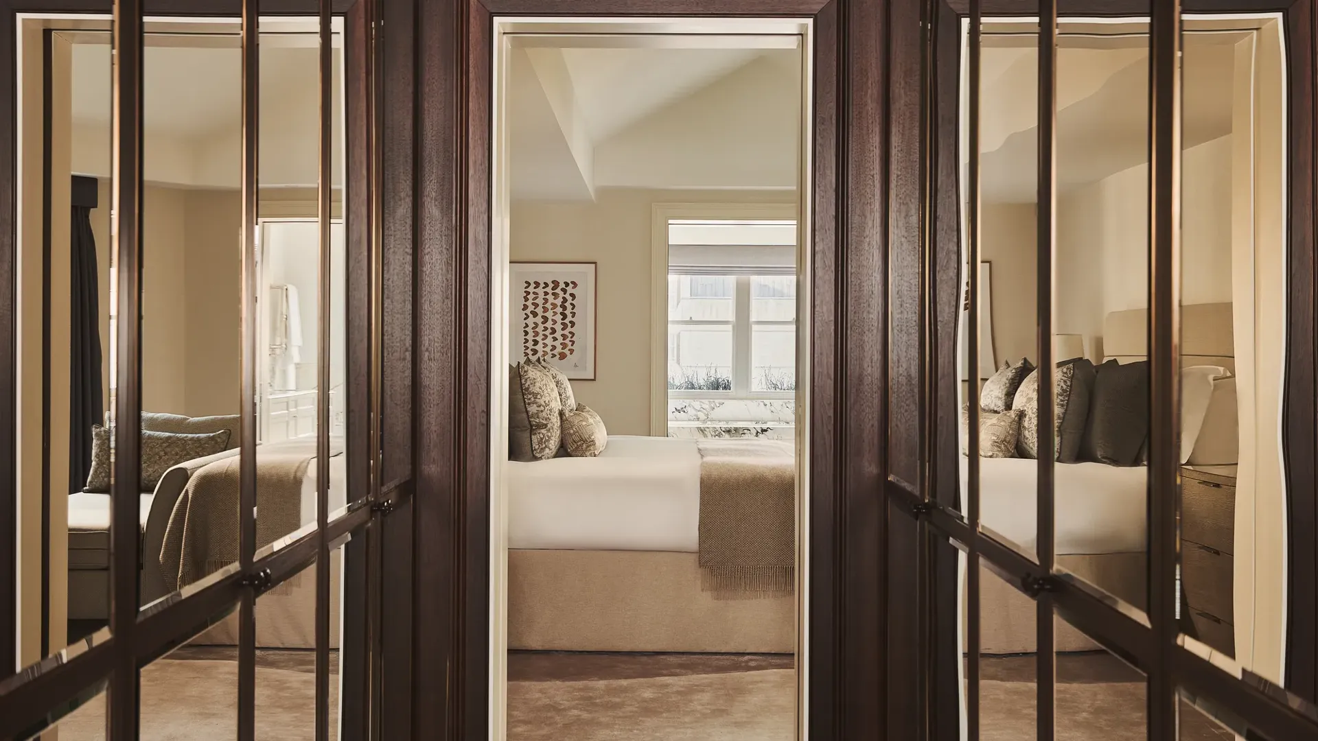 View through mirrored wardrobe doors towards a neutral-toned bedroom with a neatly made bed, soft lighting, and window bringing in natural daylight.