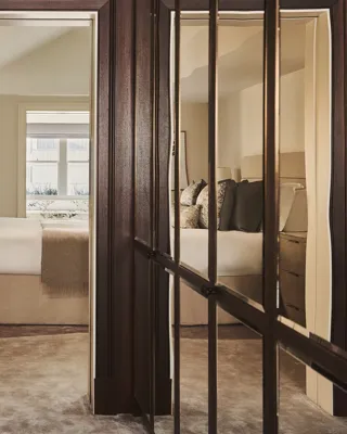 View through mirrored wardrobe doors towards a neutral-toned bedroom with a neatly made bed, soft lighting, and window bringing in natural daylight.