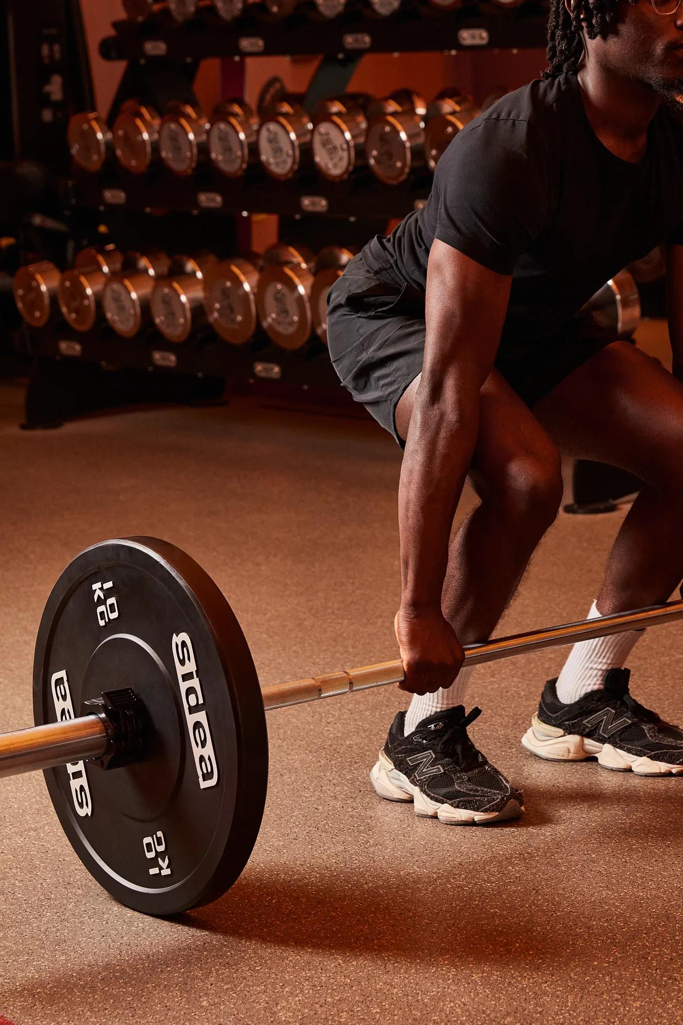 Person performing a barbell deadlift in a gym, with weight plates and dumbbells visible in the background.