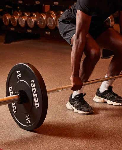 Person performing a barbell deadlift in a gym, with weight plates and dumbbells visible in the background.