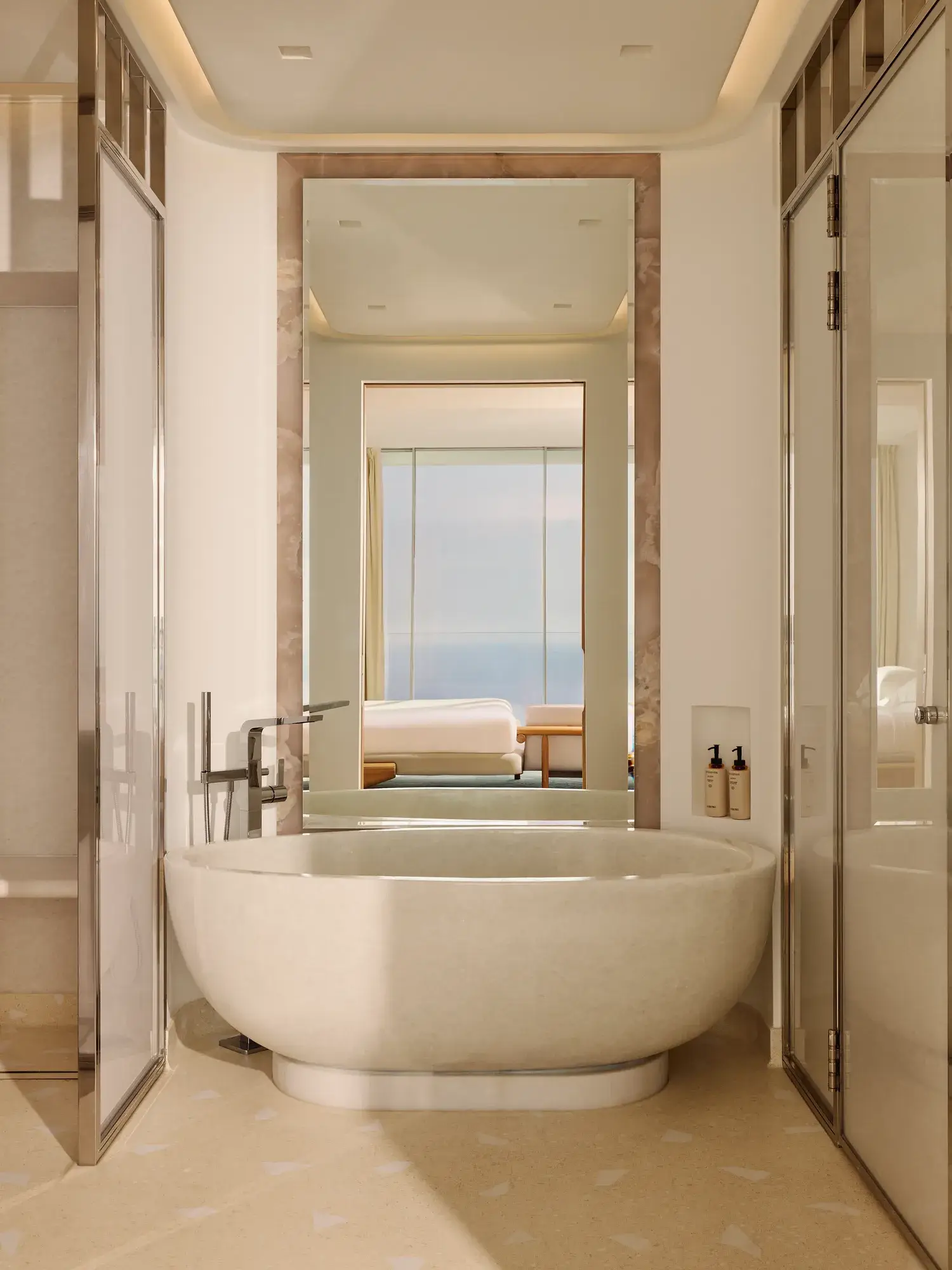 Salle de bain luxueuse avec une baignoire ovale autoportante et robinetterie moderne, grand miroir et vue sur la chambre dotée de baies vitrées offrant un panorama sur la mer.