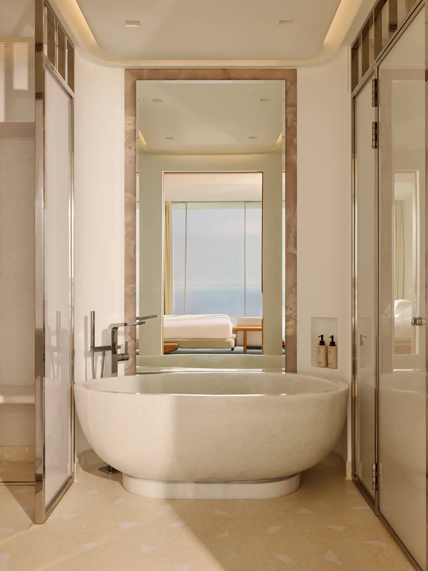 Salle de bain luxueuse avec une baignoire ovale autoportante et robinetterie moderne, grand miroir et vue sur la chambre dotée de baies vitrées offrant un panorama sur la mer.