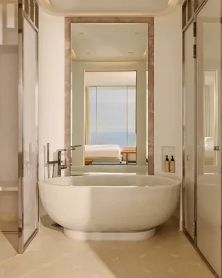 Salle de bain luxueuse avec une baignoire ovale autoportante et robinetterie moderne, grand miroir et vue sur la chambre dotée de baies vitrées offrant un panorama sur la mer.