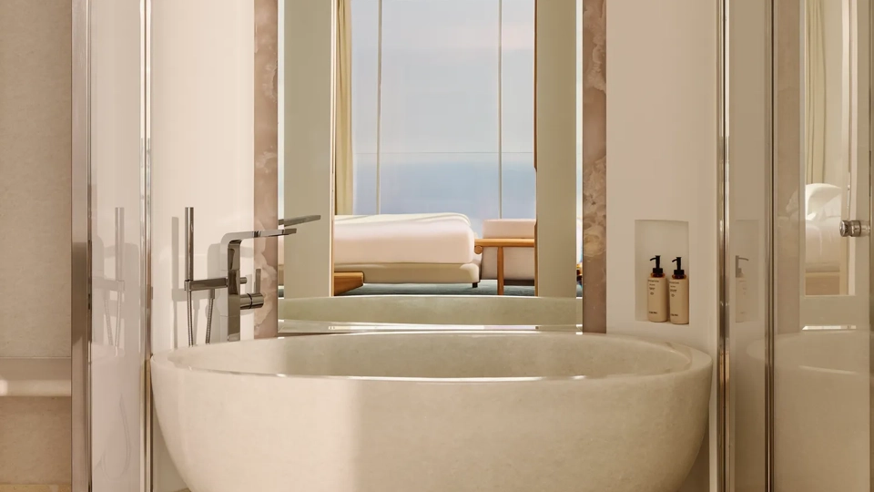 Salle de bain luxueuse avec une baignoire ovale autoportante et robinetterie moderne, grand miroir et vue sur la chambre dotée de baies vitrées offrant un panorama sur la mer.
