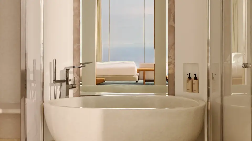 Salle de bain luxueuse avec une baignoire ovale autoportante et robinetterie moderne, grand miroir et vue sur la chambre dotée de baies vitrées offrant un panorama sur la mer.