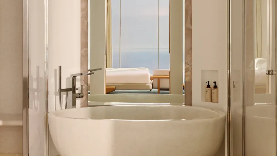 Salle de bain luxueuse avec une baignoire ovale autoportante et robinetterie moderne, grand miroir et vue sur la chambre dotée de baies vitrées offrant un panorama sur la mer.