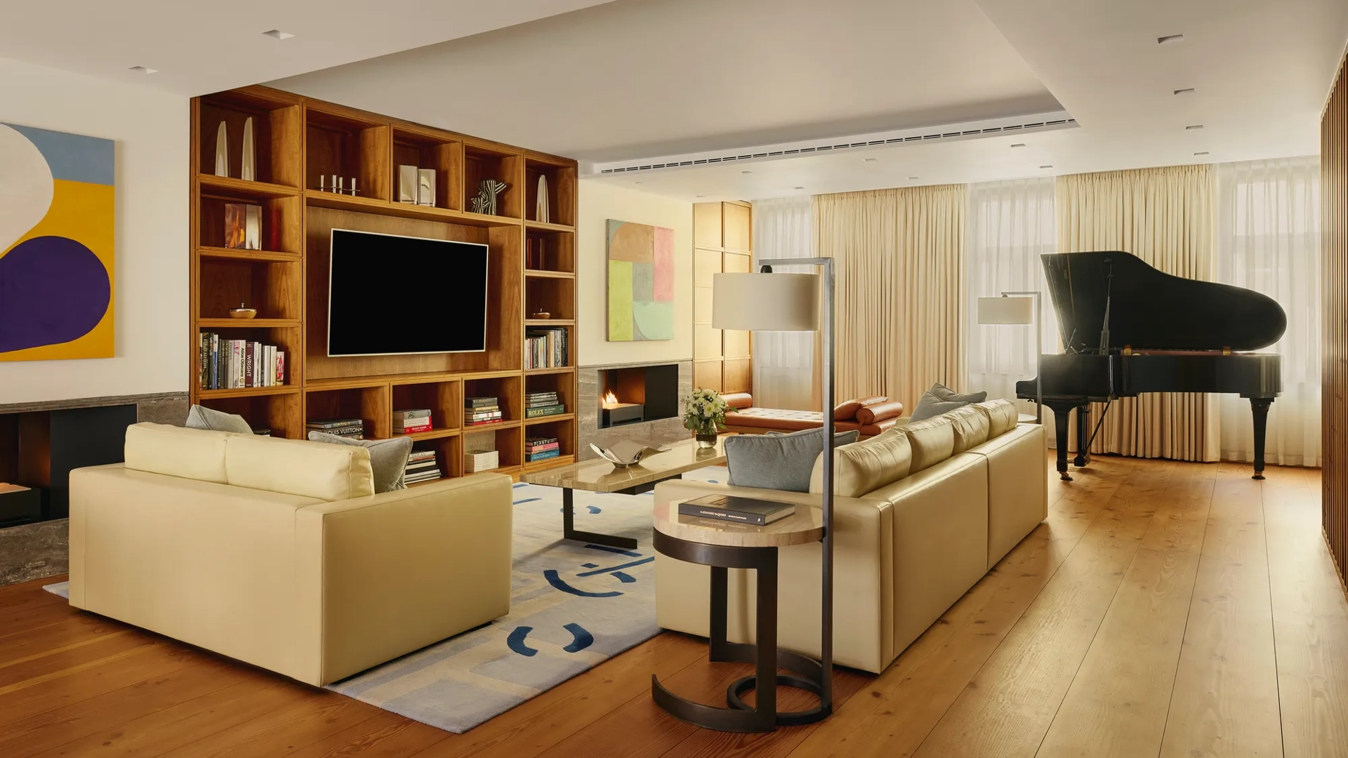 A contemporary living room with cream leather sofas, wooden flooring, built-in shelving with a large TV, abstract artwork, and a grand piano near the window, softly lit by natural light through sheer curtains.