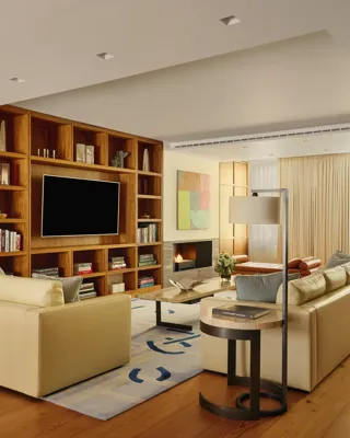 A contemporary living room with cream leather sofas, wooden flooring, built-in shelving with a large TV, abstract artwork, and a grand piano near the window, softly lit by natural light through sheer curtains.