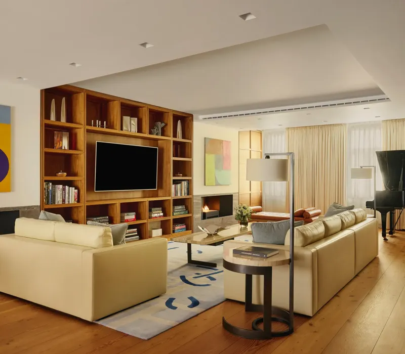 A contemporary living room with cream leather sofas, wooden flooring, built-in shelving with a large TV, abstract artwork, and a grand piano near the window, softly lit by natural light through sheer curtains.