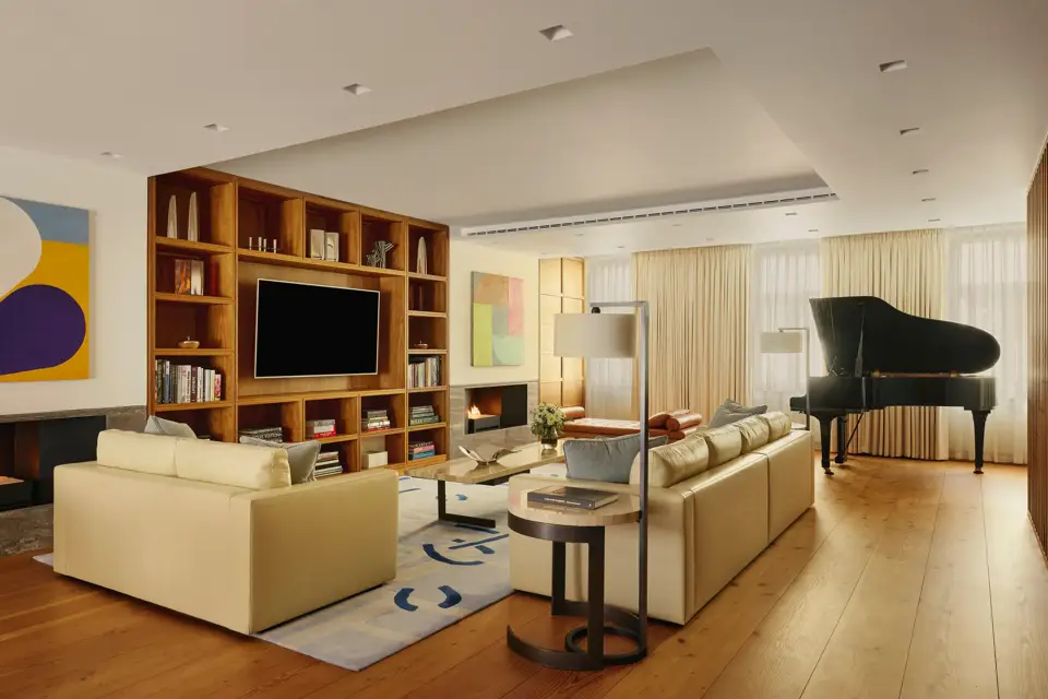 A contemporary living room with cream leather sofas, wooden flooring, built-in shelving with a large TV, abstract artwork, and a grand piano near the window, softly lit by natural light through sheer curtains.