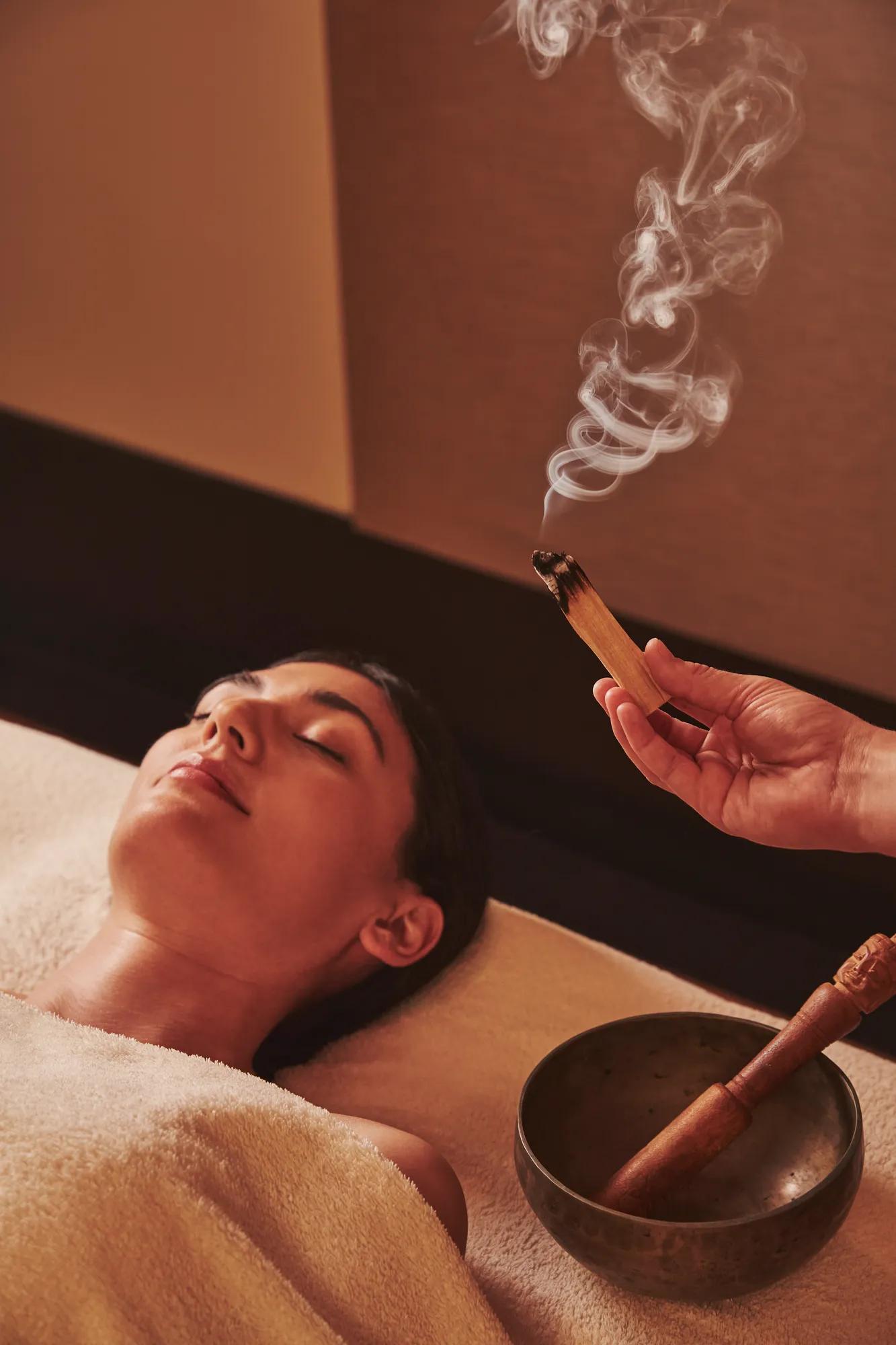 Person resting during a spa treatment while a therapist wafts scented smoke above them, with a singing bowl nearby.