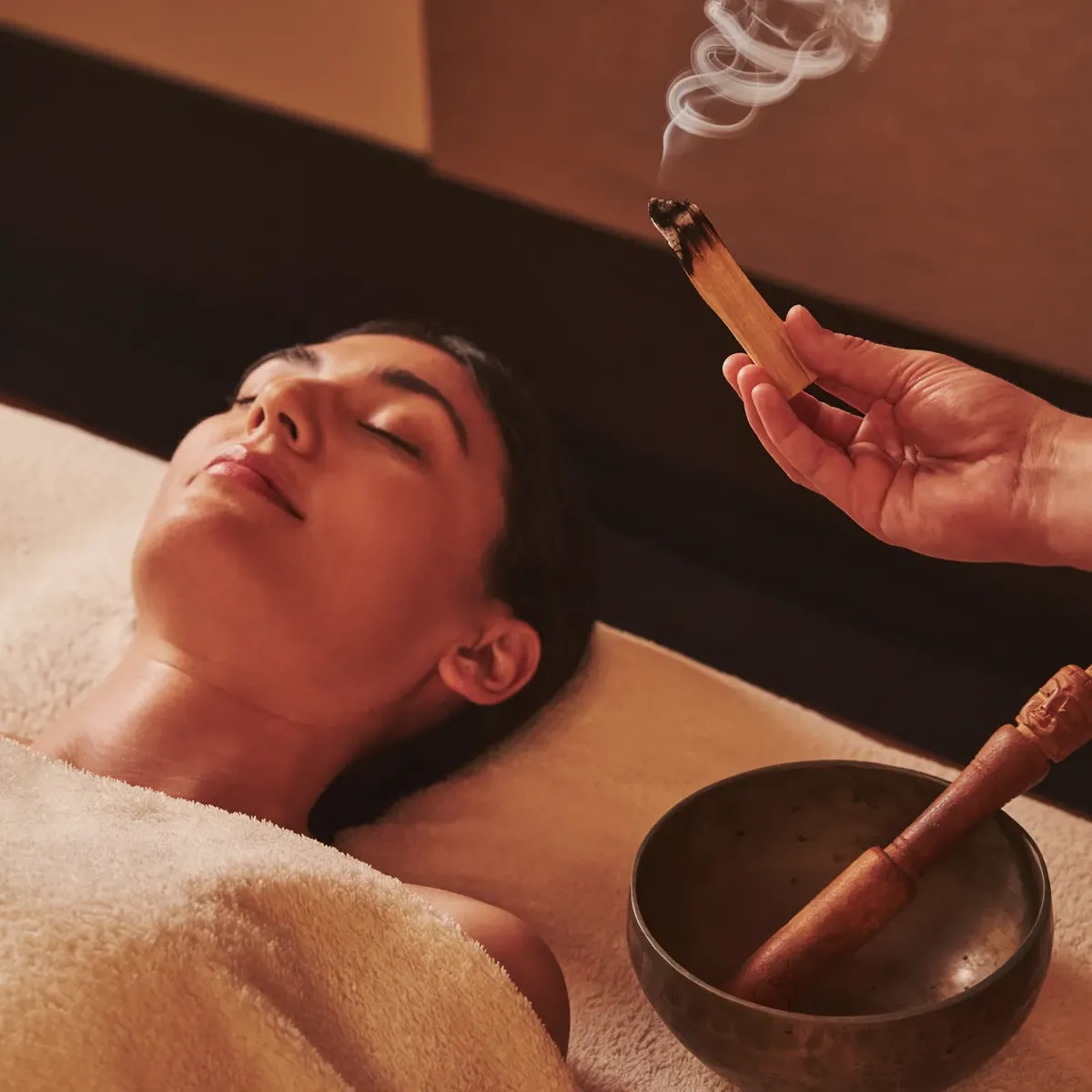 Person resting during a spa treatment while a therapist wafts scented smoke above them, with a singing bowl nearby.