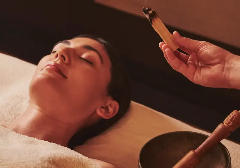 Person resting during a spa treatment while a therapist wafts scented smoke above them, with a singing bowl nearby.