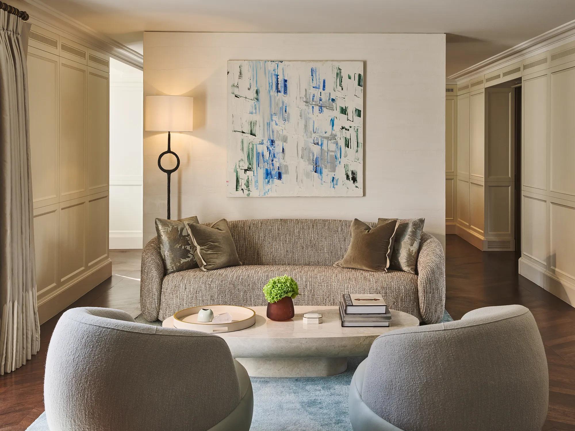 Contemporary living room with sofa, armchairs and coffee table, featuring abstract artwork, soft lighting, neutral tones, and decorative objects arranged neatly.