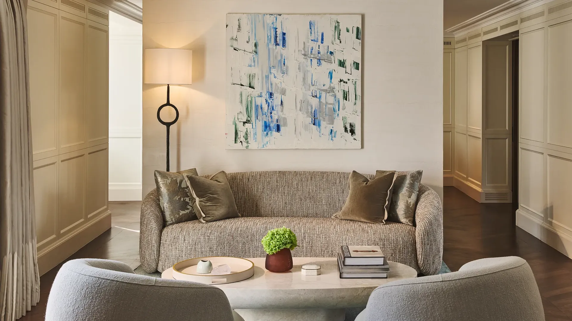Contemporary living room with sofa, armchairs and coffee table, featuring abstract artwork, soft lighting, neutral tones, and decorative objects arranged neatly.