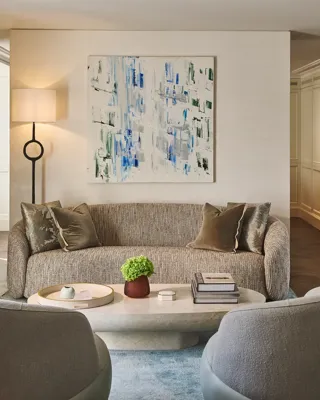Contemporary living room with sofa, armchairs and coffee table, featuring abstract artwork, soft lighting, neutral tones, and decorative objects arranged neatly.
