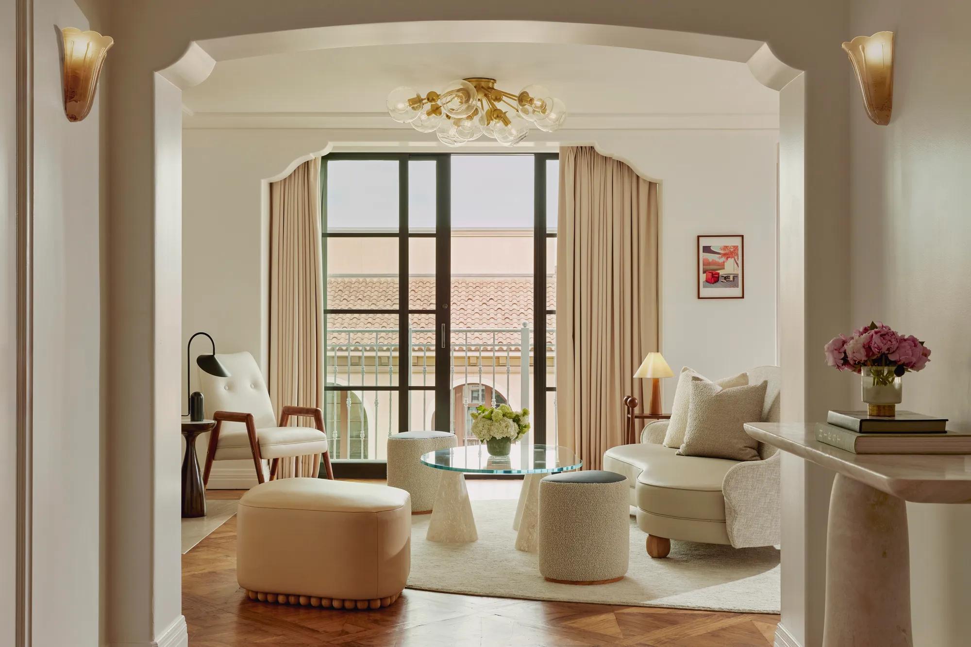Intimate sitting room facing balcony doors; glass coffee table on conical bases, boucle stools, mid‑century chair and loveseat; terracotta roofline visible outside.