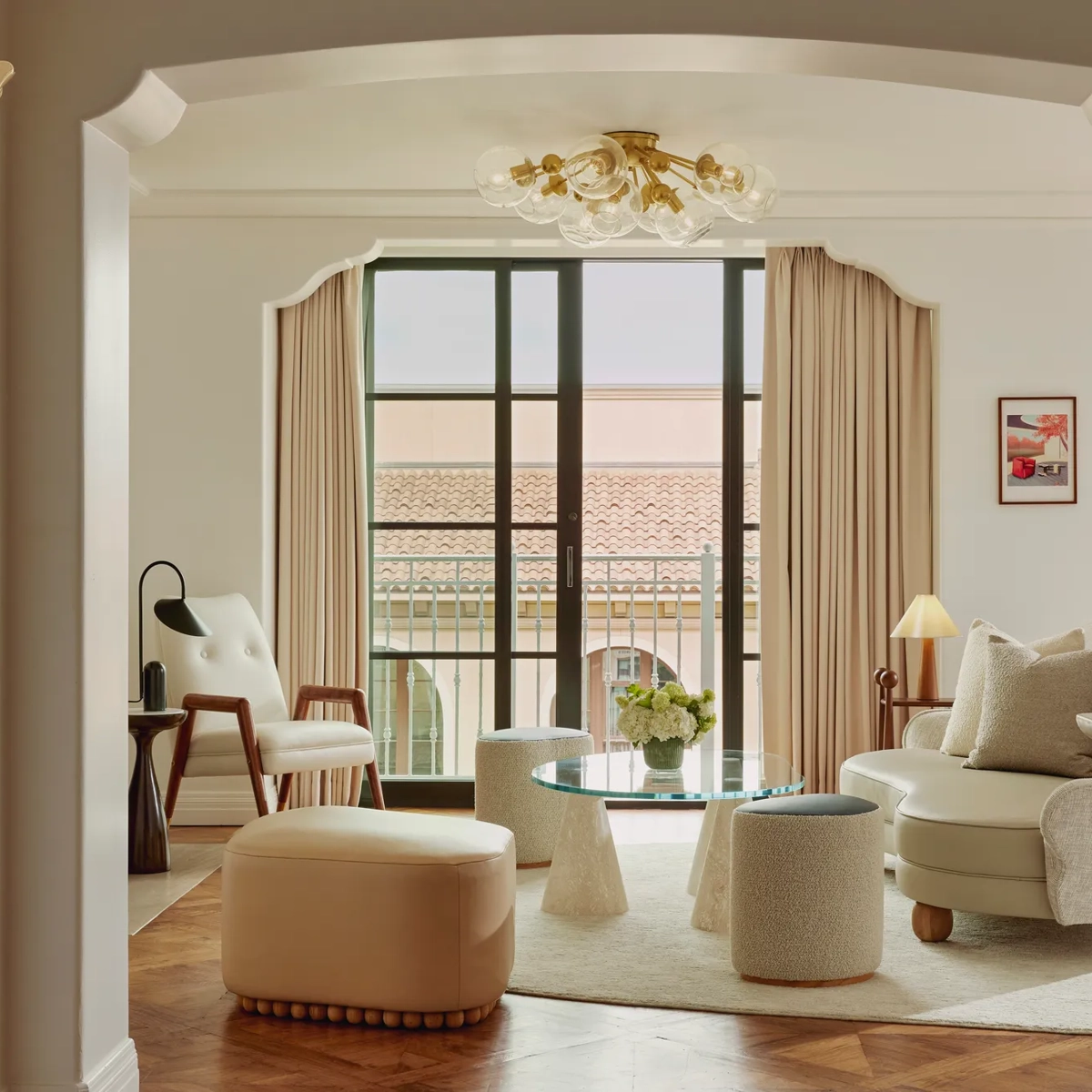 Intimate sitting room facing balcony doors; glass coffee table on conical bases, boucle stools, mid‑century chair and loveseat; terracotta roofline visible outside.
