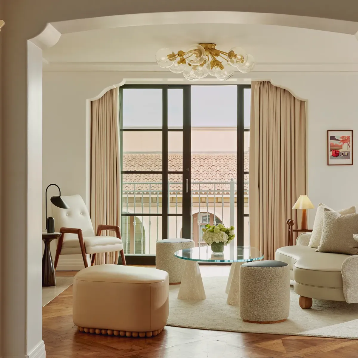 Intimate sitting room facing balcony doors; glass coffee table on conical bases, boucle stools, mid‑century chair and loveseat; terracotta roofline visible outside.