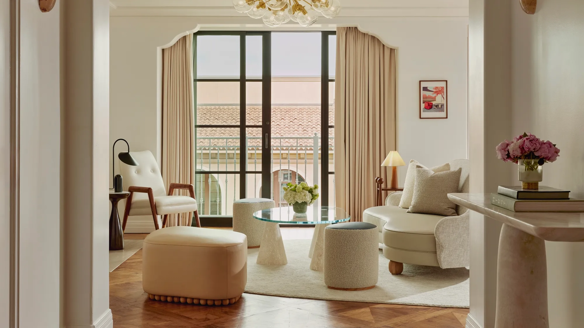 Intimate sitting room facing balcony doors; glass coffee table on conical bases, boucle stools, mid‑century chair and loveseat; terracotta roofline visible outside.