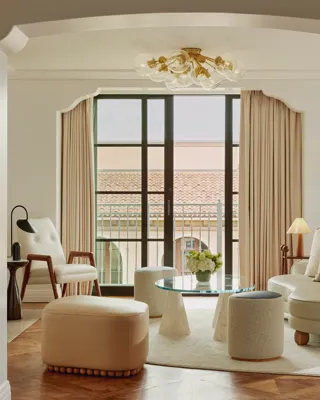 Intimate sitting room facing balcony doors; glass coffee table on conical bases, boucle stools, mid‑century chair and loveseat; terracotta roofline visible outside.