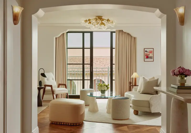 Intimate sitting room facing balcony doors; glass coffee table on conical bases, boucle stools, mid‑century chair and loveseat; terracotta roofline visible outside.